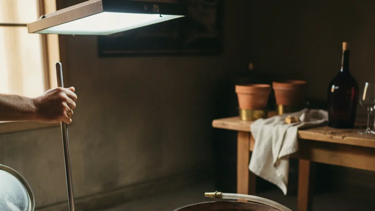 Hand stirring wine in a tank beside an oak barrel and siphon